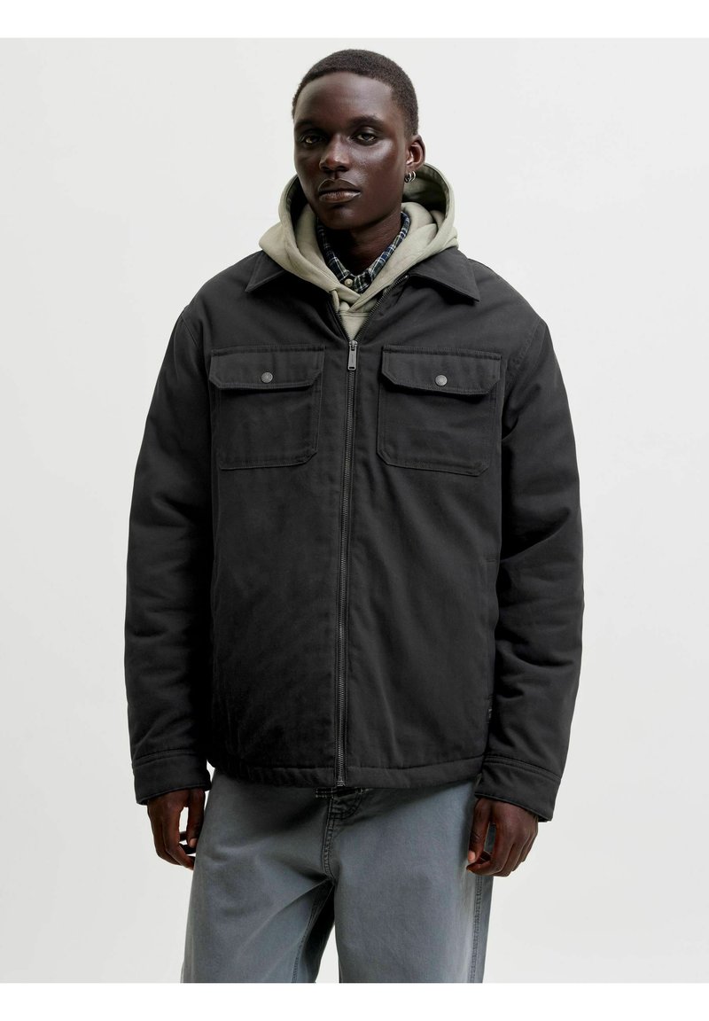 Jeune personne portant une veste noire zippée sur un sweat à capuche beige et une chemise à carreaux, debout devant un fond clair uni.