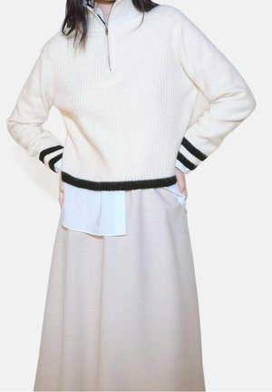 Personne portant un pull blanc à fermeture éclair avec des rayures noires sur les poignets et l'ourlet, par-dessus une chemise bleu clair et une jupe beige. Mains sur les hanches.