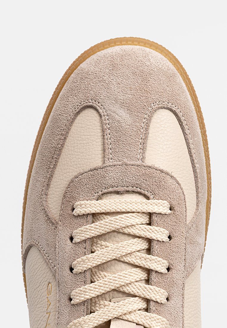 Beige Wildleder- und Leder-Sneaker mit texturierter Oberfläche, ausgestattet mit cremefarbenen Schnürsenkeln und einer hellbraunen Gummisohle. Goldenes Logo an der Seite.