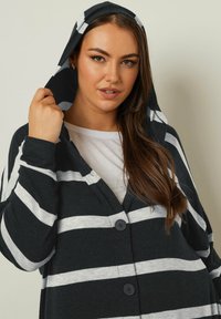 Mustat ja valkoiset raidalliset neuletakki hupulla, jossa on nappisuljenta ja pehmeä kangas. Alla näkyy valkoinen T-paita.