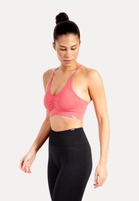 Kvinna med hår i knut som bär en rosa sport-bh och svarta leggings med hög midja, tittar neråt och lite åt sidan mot en vit bakgrund.