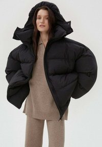 Černá péřová bunda s oversized designem, která má velkou kapuci, prošívanou strukturu a výrazné rukávy. Nosená přes béžový pletený outfit.