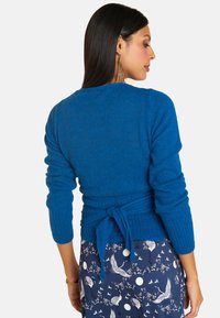 Blauer gestrickter Pullover mit einer Rückenschnürung, langen Ärmeln, gerippter Textur und einfarbigem Design. Kombiniert mit einem gemusterten Rock, der Kraniche zeigt.