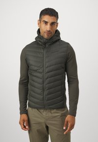 Dikke, gevoerde vest in donkergroen, gemaakt van gewatteerd materiaal met een ritssluiting aan de voorkant. Gedragen over een lange mouwen shirt en gecombineerd met lichte broeken.