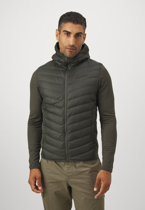 Puffy, hooded vest in dark green, made of quilted material with a zippered front. Worn over a long-sleeve shirt and paired with light pants.