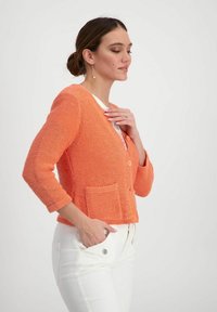 Korallenfarbener Strickcardigan mit 3/4-Ärmeln, vorderem Knopfverschluss und zwei Taschen. Getragen über einem weißen Oberteil und kombiniert mit weißen Hosen.