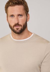 Maglione beIGE con collo rotondo e finiture testurizzate, indossato sopra una camicia bianca. Il tessuto appare morbido e ha un design aderente.
