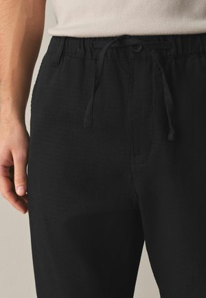 Man wearing black drawstring pants and a white shirt, showing the waistband and hand resting beside the pants.