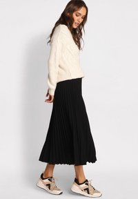 Cremefarvet kabelstriktrøje, sort plisseret midi-nederdel og beige sneakers med sorte accenter. Outfittet har en afslappet pasform og flydende form.