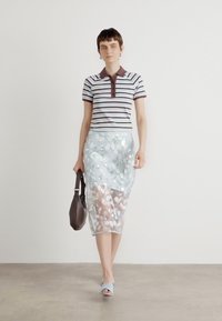Polo à rayures avec un col marron, associé à une jupe bleue transparente ornée de fleurs. Fond blanc, le mannequin porte un sac marron.