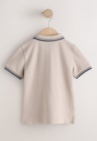 Beige short-sleeve polo shirt with navy and white striped collar and sleeve cuffs hanging on wooden hanger against white wall.