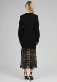 Pull en tricot noir à manches longues, associé à une jupe maxi en dentelle noire avec des détails en motifs. Des talons noirs à lanières complètent le look.