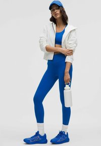 Mėlyna sportinė apranga, kurią sudaro trumpas viršus, prigludę legginiai ir mėlyni sportbačiai. Aprangą papildo balti vėjara ir balta vandens buteliukas.