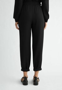 Kobieta w czarnych, wysokich, skróconych spodniach z podwiniętymi nogawkami oraz czarnych sandałach z odkrytymi palcami, stojąca na tle jednolitego jasnego tła.