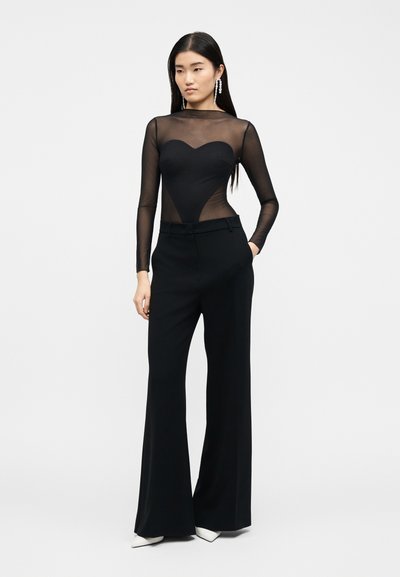 Γυναίκα που φοράει μαύρο διαφανές μακρυμάνικο τοπ με αδιάφανο μπούστο σε σχήμα καρδιάς, μαύρο παντελόνι με φαρδιά πόδια, λευκά μυτερά παπούτσια και κρεμαστά σκουλαρίκια.