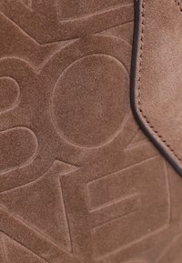 Sac en daim marron avec un motif de texte embossé, présentant une texture lisse et des coutures sombres contrastées le long du bord.