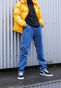 Giacca puffer gialla sopra un fleece nero, jeans in denim blu e scarpe nere con dettagli bianchi. In piedi contro uno sfondo ondulato.