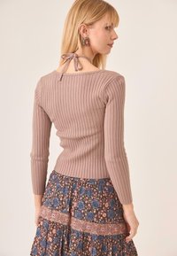 Mujer rubia con una blusa de manga larga de canalé en tono malva y un escote tipo halter atado, combinada con una falda de estampado floral en tonos púrpura y azul.