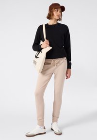 Top negro de manga larga, joggers beige con cordón, zapatillas blancas y gorra marrón. El modelo lleva una bolsa de tote beige con un logo negro.