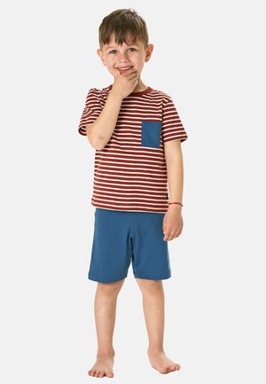 Young boy standing barefoot, wearing a red and white striped shirt with a blue pocket and blue shorts, smiling and touching his chin.