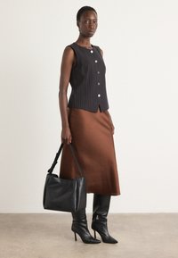 Top senza maniche a righe nere con bottoni tondi, abbinato a una gonna di raso marrone. La modella tiene una borsa in pelle nera, indossando stivali neri eleganti.