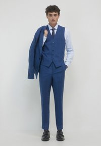 Blau karierter Dreiteiler mit taillierter Passform, bestehend aus einer Weste, einer Jacke mit zwei Knöpfen und schmalen Hosen. Schwarze Schuhe vervollständigen den Look.