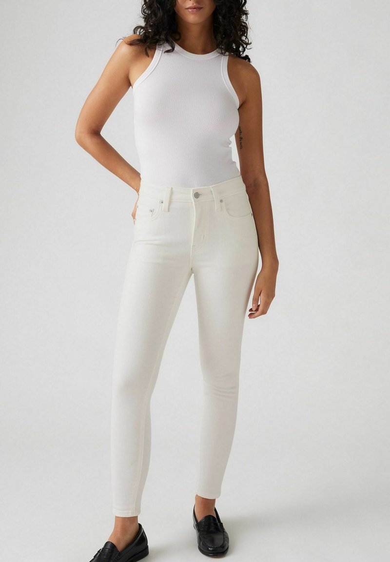 Camiseta de tirantes blanca de canalé combinada con unos jeans blancos ajustados. Los jeans presentan una textura suave y un diseño clásico de cinco bolsillos. Unos mocasines negros completan el look.