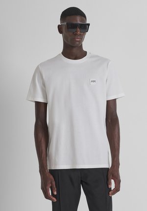 Witte katoenen T-shirt met een ronde hals, korte mouwen en een klein vierkant logo op de linkerborst, gedragen met donkere broeken en een zonnebril.