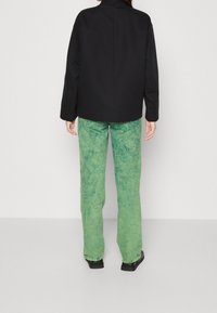 Zwarte oversized jas met een hoge kraag, gecombineerd met lichtgroene, textuurloze, loszittende broeken. Zwarte, platte schoenen maken de look compleet.