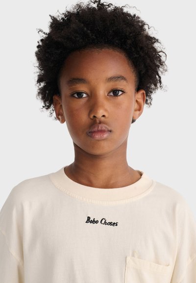 Barn med krøllet hår iført en cremefarvet sweatshirt med "Bobo Choses" broderet på brystet, kigger lige frem.