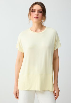 Femme portant un haut ample à manches courtes jaune pâle avec un pantalon blanc, debout devant un fond uni gris clair.
