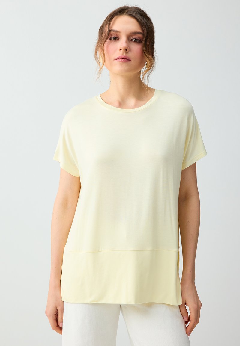 Femme portant un haut ample à manches courtes jaune pâle avec un pantalon blanc, debout devant un fond uni gris clair.