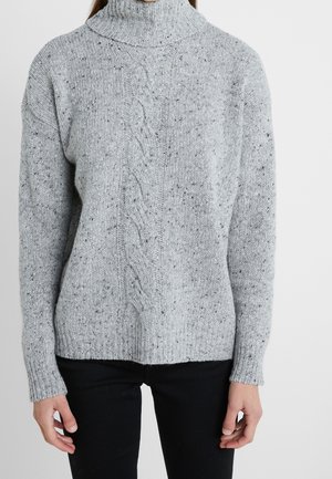 Personne portant un pull à col roulé gris clair en maille torsadée avec des taches noires et un pantalon noir, debout devant un fond blanc uni.