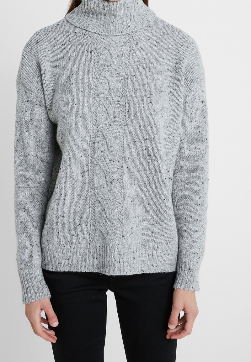 Personne portant un pull à col roulé gris clair en maille torsadée avec des taches noires et un pantalon noir, debout devant un fond blanc uni.