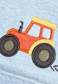 Stickerei eines roten Traktors auf einem hellblauen Stoff, mit runden schwarzen Rädern und gelben Akzenten an der Kabine und dem Dach.