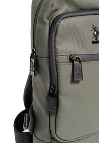 Zielona oliwkowa plecak wykonany z teksturowanej tkaniny z czarnymi akcentami, dwoma zamykanymi kieszeniami i logo umieszczonym z przodu.