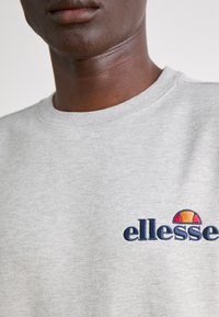 Sudadera de color gris claro hecha de algodón, con un cuello redondo acanalado y el logotipo bordado de "ellesse" en color marino, acompañado de un gráfico de sol colorido.