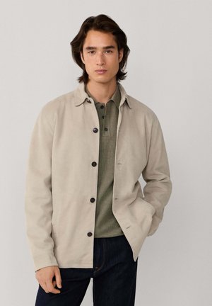 Joven con una chaqueta beige abotonada sobre una camisa polo verde oliva y vaqueros oscuros, de pie con una mano en el bolsillo.