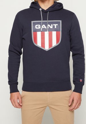 Persona con sudadera con capucha azul marino con logo de escudo GANT en rojo, blanco y gris en el pecho, combinada con pantalones beige.