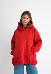 Rode oversized hoodie gemaakt van zachte stof, met een grote voorzak, geribbelde manchetten en een capuchon met trekkoord. Draagt bij lichte blauwe jeans.