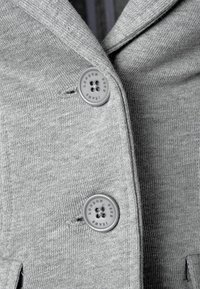 Grått stickat tygblazer med två runda grå knappar, varav en är graverad med "LONDON", samt en snedskuren krage. Slät textur och lättviktsdesign.