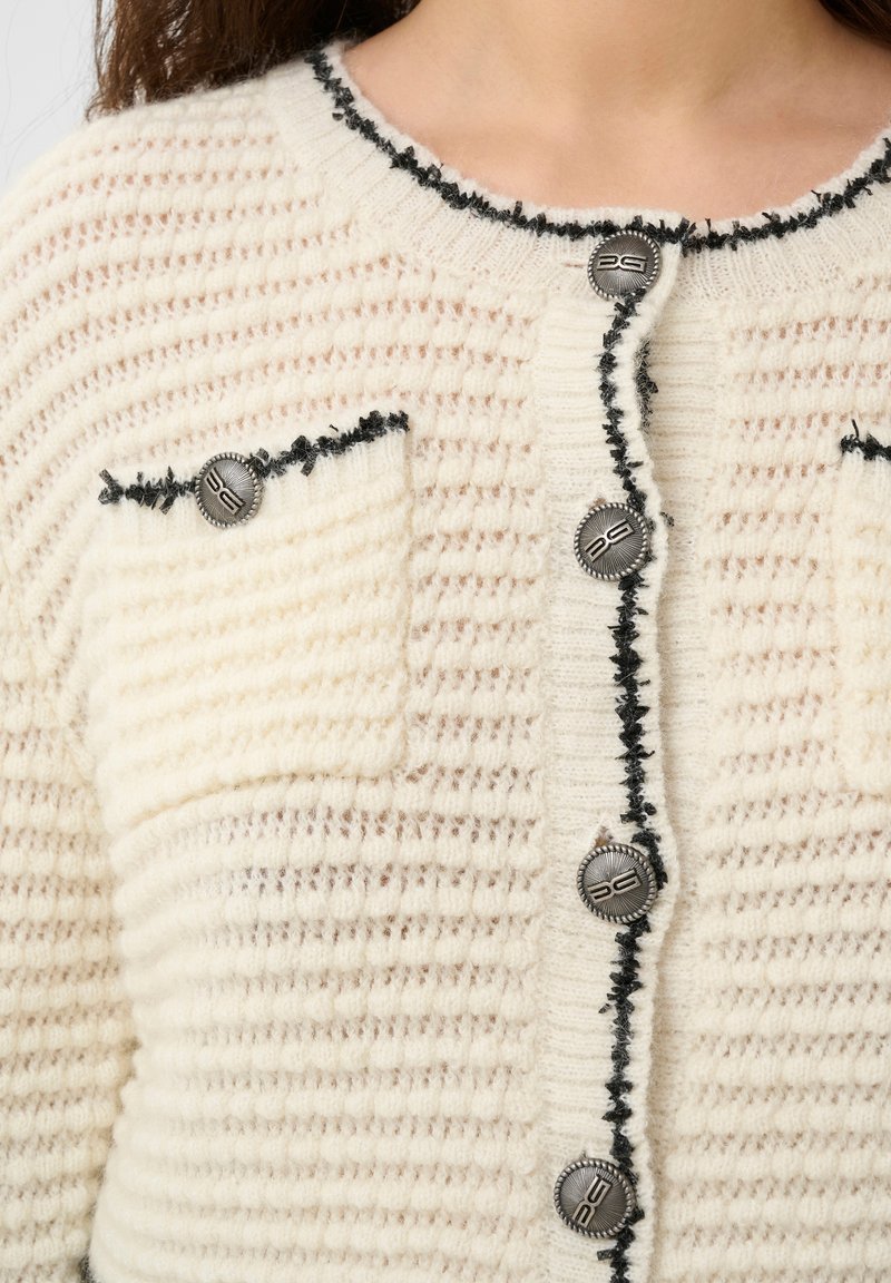 Cremfarbener Cardigan mit einem strukturierten Muster, schwarzem Besatz entlang der Kanten, einer Brusttasche und silbernen Knöpfen mit Logo-Detail.