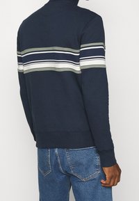 Muž v námořnickém modrém svetru s bílými a šedými vodorovnými pruhy na zádech a horních rukávech, v kombinaci s modrými džíny.