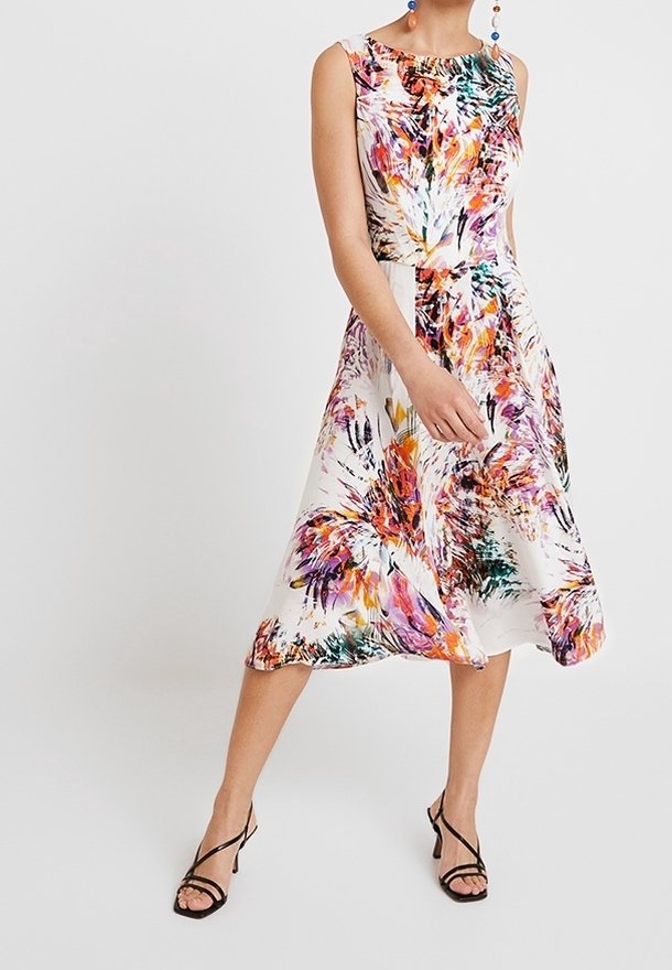Femme portant une robe midi sans manches à fleurs multicolores avec des sandales noires à talons hauts à brides, debout devant un fond uni.