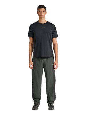 Młody mężczyzna stoi, ubrany w czarną sportową koszulkę z krótkim rękawem oraz ciemnoszare spodnie dresowe z czarnymi butami sportowymi, zwrócony przodem.