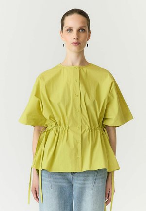 Kvinde med håret sat tilbage iført limegrøn kortærmet skjorte med knapper og snøre i taljen samt lyseblå jeans mod en ensfarvet baggrund.