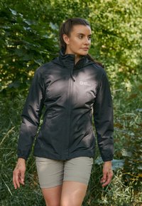 Veste noire, légère et imperméable, avec capuche et logo, présentant une coupe ajustée et une texture lisse, portée sur un short beige dans un environnement verdoyant.