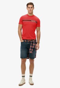Rode t-shirt met korte mouwen, horizontale strepen en logo, gecombineerd met denimshorts en een geruite blouse om de taille geknoopt. Witte en bruine sneakers.