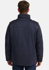 Marineblauwe, gevoerde jas met een hoge kraag, schouderepauletten en zijzakken. De stof lijkt glad met een matte afwerking.