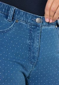 Vaqueros de mezclilla azul con un acabado ligeramente desvaído, que presentan un pequeño patrón de puntos blancos, un cierre de botón metálico y un bolsillo delantero plano.